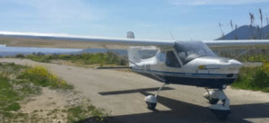 Un avión monomotor, blanco y azul, se encuentra en una pista de tierra rural en Atarfe con un majestuoso telón de fondo de montañas, invitándote a experimentar "Piloto por un día en Atarfe". - ForTwo - Planes para parejas