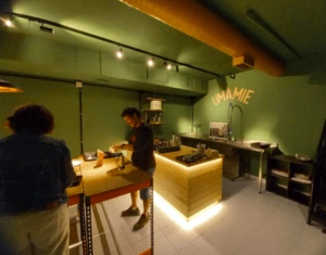 En la cocina verde de UMAMIE, dos personas trabajan en la barra y el fregadero, preparando pinchos. La escena evoca la experiencia de un Taller de Pinchos en Bilbao, reflejando la vibrante pasión que se siente en Bilbao mientras se perfeccionan juntos exquisitos bocados. - ForTwo - Planes para parejas