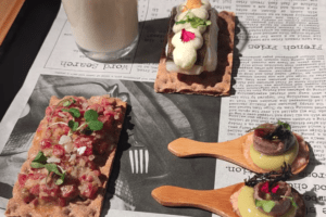 Una deliciosa variedad de aperitivos presentados en un periódico, con coberturas de galletas y bocados servidos con cuchara, combina perfectamente con una bebida cremosa, capturando los sabores vibrantes de Taller de Pinchos en Bilbao. - ForTwo - Planes para parejas