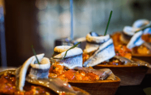 Primer plano de un plato de pescado elegantemente presentado con guarniciones y salsa, evocando una creación del famoso Taller de Pinchos de Bilbao, todo bellamente dispuesto en bandejas de madera para servir. - ForTwo - Planes para parejas