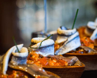 Primer plano de un plato de pescado elegantemente presentado con guarniciones y salsa, evocando una creación del famoso Taller de Pinchos de Bilbao, todo bellamente dispuesto en bandejas de madera para servir. - ForTwo - Planes para parejas
