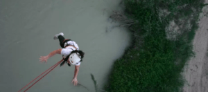 Una persona con el arnés "Puenting en el río Genil" salta sobre el río con los brazos extendidos, mientras se ven orillas cubiertas de hierba debajo. - ForTwo - Planes para parejas
