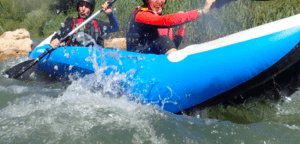 Dos personas reman en un kayak inflable azul a través de los emocionantes rápidos del Río Mijares, salpicando agua por todas partes mientras abrazan la emocionante aventura del Rafting en el Río Mijares. - ForTwo - Planes para parejas