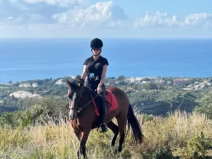 Una persona monta elegantemente a caballo por una colina cubierta de hierba con el océano y el cielo de fondo, evocando el encanto de la Ruta a Caballo en Marbella. - ForTwo - Planes para parejas