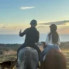 Dos personas a caballo disfrutan de una Ruta a Caballo en Marbella al atardecer, capturando una selfie con la vista panorámica del océano y su fiel perro cerca. - ForTwo - Planes para parejas