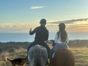 Dos personas a caballo disfrutan de una Ruta a Caballo en Marbella al atardecer, capturando una selfie con la vista panorámica del océano y su fiel perro cerca. - ForTwo - Planes para parejas