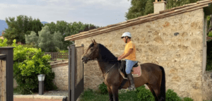 Con casco, una persona monta a caballo en la Ruta a Caballo por el Valle del Río Ser, pasando junto a una construcción de piedra en medio de la exuberante vegetación. - ForTwo - Planes para parejas