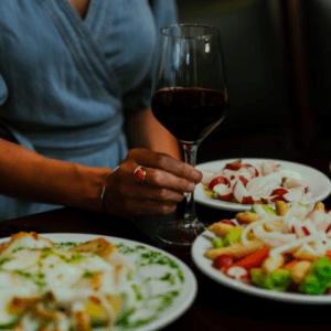 Una persona disfrutando de una copa de vino tinto en una mesa con pasta y ensalada, evocando delicias similares a las de la Ruta de Tapas y Vinos de Valencia. - ForTwo - Planes para parejas