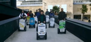 Un grupo se desliza sin esfuerzo por la Ruta en Segway por el Monte del Castro, sosteniendo carteles coloridos mientras recorren el sendero frente a un edificio hacia unas vistas impresionantes. - ForTwo - Planes para parejas