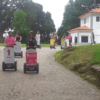 La gente que realiza la Ruta en Segway por el Monte del Castro se desliza por un camino pavimentado a través de una exuberante vegetación y árboles, con el sereno telón de fondo de un edificio blanco. - ForTwo - Planes para parejas