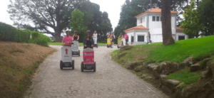 La gente que realiza la Ruta en Segway por el Monte del Castro se desliza por un camino pavimentado a través de una exuberante vegetación y árboles, con el sereno telón de fondo de un edificio blanco. - ForTwo - Planes para parejas