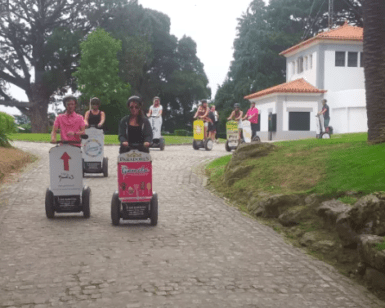 La gente que realiza la Ruta en Segway por el Monte del Castro se desliza por un camino pavimentado a través de una exuberante vegetación y árboles, con el sereno telón de fondo de un edificio blanco. - ForTwo - Planes para parejas