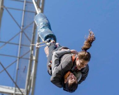 Dos personas disfrutan de un Salto Bungee Tándem en Lloret de Mar, colgando boca abajo contra un cielo azul claro. - ForTwo - Planes para parejas