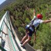 En Valencia, una persona con casco rojo y arnés experimenta la emoción del "Salto de puenting en Valencia con vídeo: ¡vive la adrenalina en pareja!" mientras salta desde un puente sobre un bosque ante la mirada de los espectadores. - ForTwo - Planes para parejas