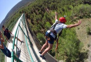 En Valencia, una persona con casco rojo y arnés experimenta la emoción del "Salto de puenting en Valencia con vídeo: ¡vive la adrenalina en pareja!" mientras salta desde un puente sobre un bosque ante la mirada de los espectadores. - ForTwo - Planes para parejas