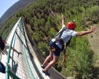 En Valencia, una persona con casco rojo y arnés experimenta la emoción del "Salto de puenting en Valencia con vídeo: ¡vive la adrenalina en pareja!" mientras salta desde un puente sobre un bosque ante la mirada de los espectadores. - ForTwo - Planes para parejas