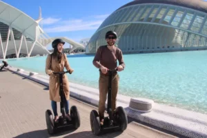 Dos personas viajan en Segway por la Valencia medieval, con cascos y sonriendo junto a un edificio futurista y una piscina, mostrando la mezcla de arquitectura antigua y nueva. - ForTwo - Planes para parejas