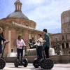 Gente disfrutando de un recorrido "Segway por la Valencia medieval" cerca de una fuente en una plaza histórica de Valencia, rodeada de ornamentados edificios medievales y una gran estructura abovedada. - ForTwo - Planes para parejas