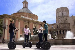 Gente disfrutando de un recorrido "Segway por la Valencia medieval" cerca de una fuente en una plaza histórica de Valencia, rodeada de ornamentados edificios medievales y una gran estructura abovedada. - ForTwo - Planes para parejas
