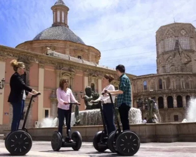 Gente disfrutando de un recorrido "Segway por la Valencia medieval" cerca de una fuente en una plaza histórica de Valencia, rodeada de ornamentados edificios medievales y una gran estructura abovedada. - ForTwo - Planes para parejas