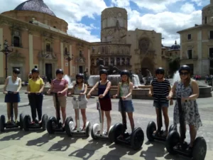 Un grupo de personas en el tour "Segway por la Valencia medieval" se desliza por una plaza medieval europea en Valencia, rodeada de edificios históricos y una pintoresca fuente. - ForTwo - Planes para parejas