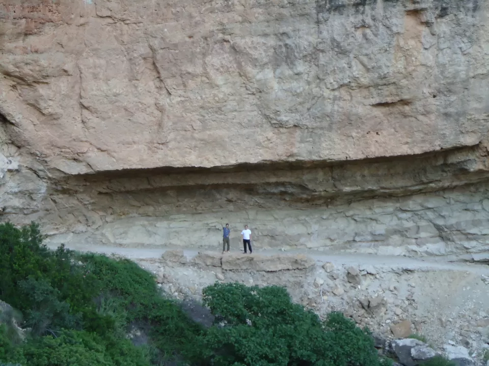 En Cerro de Agras, dos personas recorren un estrecho sendero al borde de un acantilado debajo de una imponente pared de roca, con una exuberante vegetación debajo, un lugar ideal para el senderismo por el volcán Cerro de Agras entre los restos de antiguas formaciones volcánicas. - ForTwo - Planes para parejas