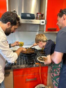 En una cocina bulliciosa, un chef formado por Michelin dirige una "Sesión de paella en casa de un chef con formación Michelin", cocinando expertamente con la ayuda de un hombre y un niño, mientras otro hombre ayuda. - ForTwo - Planes para parejas