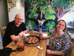 Dos personas sonrientes en una mesa con comida y bebida, incluida una deliciosa Sesión de paella en casa de un chef con formación Michelin, con un vibrante mural de la jungla de fondo. - ForTwo - Planes para parejas