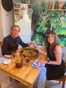 Una pareja disfruta de la "Sesión de paella en casa de un chef con formación Michelin" en una mesa de comedor de madera, con bebidas y un vibrante mural de plantas de fondo. - ForTwo - Planes para parejas