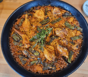 La "Sesión de paella en casa de un chef con formación Michelin" incluye una sartén bellamente decorada con pollo, arroz y verduras frescas con romero, presentada elegantemente sobre una mesa de madera. - ForTwo - Planes para parejas