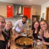 Cinco personas sonríen y posan alrededor de una gran paella en una acogedora cocina, capturando la alegría de una "Sesión de paella en casa de un chef con formación Michelin". - ForTwo - Planes para parejas