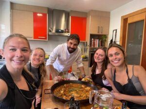 Cinco personas sonríen y posan alrededor de una gran paella en una acogedora cocina, capturando la alegría de una "Sesión de paella en casa de un chef con formación Michelin". - ForTwo - Planes para parejas