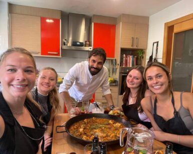 Cinco personas sonríen y posan alrededor de una gran paella en una acogedora cocina, capturando la alegría de una "Sesión de paella en casa de un chef con formación Michelin". - ForTwo - Planes para parejas