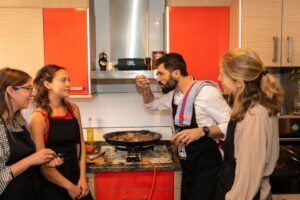 En una acogedora cocina, un hombre con formación de chef Michelin prueba la paella del fuego mientras tres mujeres miran y sonríen, disfrutando de esta "Sesión de paella en casa de un chef con formación Michelin". - ForTwo - Planes para parejas