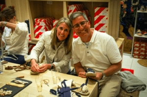 Dos personas sonríen mientras elaboran zapatos en una mesa de madera, inmersos en la atmósfera vibrante de un Taller de Alpargatas en Valencia. - ForTwo - Planes para parejas