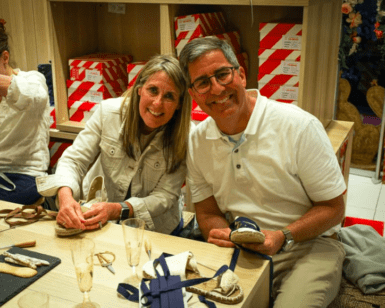 Dos personas sonríen mientras elaboran zapatos en una mesa de madera, inmersos en la atmósfera vibrante de un Taller de Alpargatas en Valencia. - ForTwo - Planes para parejas