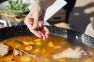 Unas manos elegantes rocían azafrán en una sartén con sabrosa carne, verduras vibrantes y un caldo rico, creando un plato que recuerda a la experiencia completa del Taller de Paella en Villa Indiano. - ForTwo - Planes para parejas