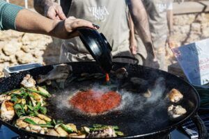 En Villa Indiano, disfruta de la "Experiencia Completa del Taller de Paella" añadiendo salsa a una gran sartén de paella con verduras y carne cocinándose al aire libre. - ForTwo - Planes para parejas