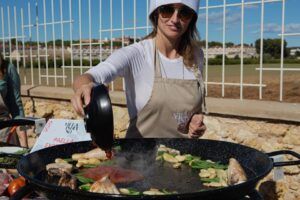 Un chef del Taller de Paella Full Experience en Villa Indiano cocina hábilmente una deliciosa paella al aire libre en una sartén grande, con un sombrero y delantal blancos frente a un encantador fondo rústico. - ForTwo - Planes para parejas