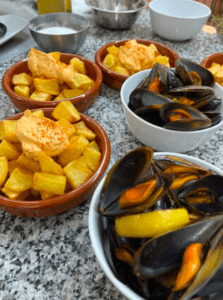 Cuencos de patatas bravas con salsa y cuencos de mejillones al vapor se sientan invitadoramente en la mesa de granito, evocando un sabor que recuerda a la experiencia del Taller de Paella y Sangría en Valencia. - ForTwo - Planes para parejas