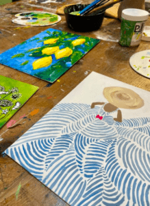 Las pinturas sobre una mesa de madera muestran limones, un diseño estampado y una figura con sombrero, con materiales de arte que hacen alusión al Taller de Pintura Acrílica en Palma. - ForTwo - Planes para parejas