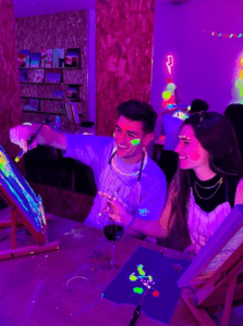 En una fiesta vibrante, dos personas pintan alegremente con colores neón bajo luz negra. La escena personifica el Taller de Pintura y Vino, donde el arte se fusiona con la celebración, y todos sonríen como si hubieran encontrado la combinación perfecta de creatividad y festividad. - ForTwo - Planes para parejas