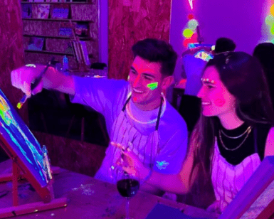 En una fiesta vibrante, dos personas pintan alegremente con colores neón bajo luz negra. La escena personifica el Taller de Pintura y Vino, donde el arte se fusiona con la celebración, y todos sonríen como si hubieran encontrado la combinación perfecta de creatividad y festividad. - ForTwo - Planes para parejas