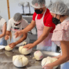 En una cocina llena de actividad, trabajadores con redecilla y mascarilla dan forma diligentemente a las bolas de masa, llenando el aire con el rico aroma del Taller de Queso Mahón-Menorca en Ferreries. - ForTwo - Planes para parejas
