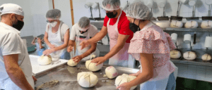 En una cocina llena de actividad, trabajadores con redecilla y mascarilla dan forma diligentemente a las bolas de masa, llenando el aire con el rico aroma del Taller de Queso Mahón-Menorca en Ferreries. - ForTwo - Planes para parejas