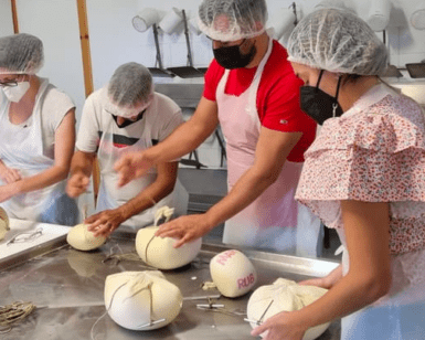 En una cocina llena de actividad, trabajadores con redecilla y mascarilla dan forma diligentemente a las bolas de masa, llenando el aire con el rico aroma del Taller de Queso Mahón-Menorca en Ferreries. - ForTwo - Planes para parejas