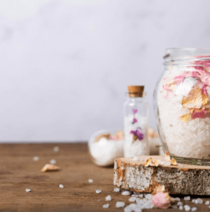 Un frasco de Taller Sales de baño mágico en Mallorca con pétalos rosas reposa sobre una superficie de madera, capturando el encanto sereno de Mallorca contra un fondo suave y neutro. - ForTwo - Planes para parejas