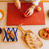 Una persona pica con destreza un tomate sobre una tabla de cortar roja entre pan, frutas y especias, encarnando la esencia vibrante de una cocina mallorquina como la que se ve en el "Taller de cocina mediterránea en Mallorca". - ForTwo - Planes para parejas