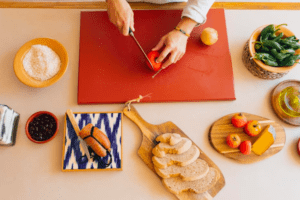 Una persona pica con destreza un tomate sobre una tabla de cortar roja entre pan, frutas y especias, encarnando la esencia vibrante de una cocina mallorquina como la que se ve en el "Taller de cocina mediterránea en Mallorca". - ForTwo - Planes para parejas