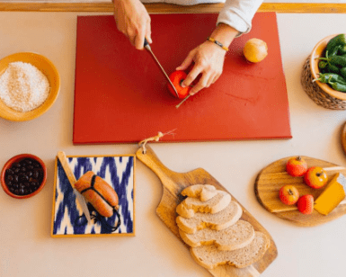 Una persona pica con destreza un tomate sobre una tabla de cortar roja entre pan, frutas y especias, encarnando la esencia vibrante de una cocina mallorquina como la que se ve en el "Taller de cocina mediterránea en Mallorca". - ForTwo - Planes para parejas